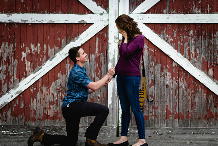 Man playfully proposing on one knee while girlfriend reacts with surprise in front of rustic red barn doors Man playfully proposing on one knee while girlfriend reacts with surprise in front of rustic red barn doors