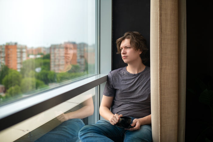 Young man sitting by the window, reflecting thoughtfully, illustrating the impact of helicopter parents on relationships. Young man sitting by the window, reflecting thoughtfully, illustrating the impact of helicopter parents on relationships.