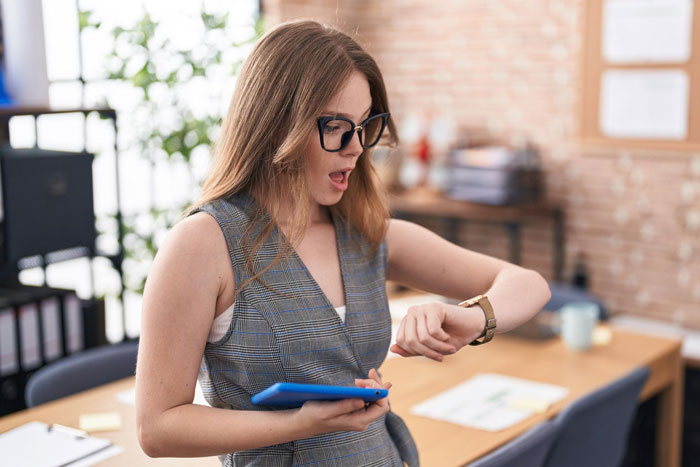 Young female employee looking shocked at wristwatch in office, depicting punctuality obsession and clever workaround at work. Young female employee looking shocked at wristwatch in office, depicting punctuality obsession and clever workaround at work.