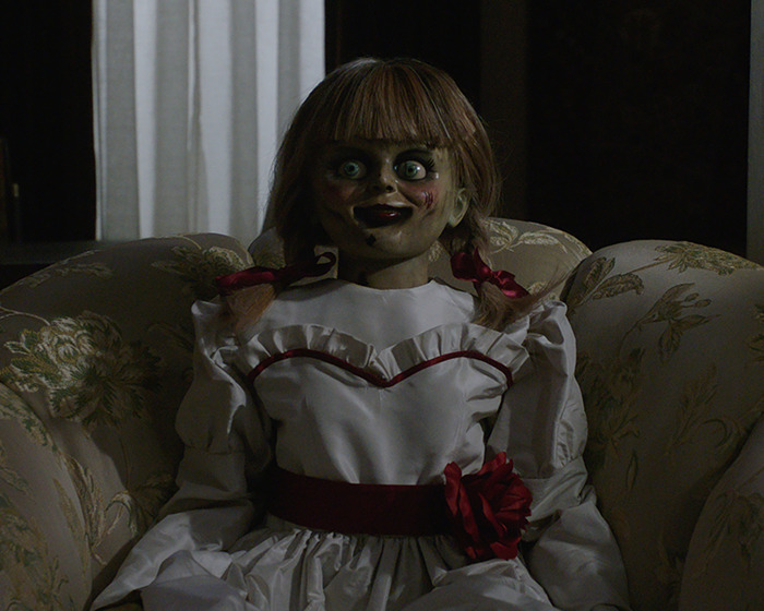 Haunted Annabelle doll with eerie smile sitting in a vintage armchair in a dimly lit room.