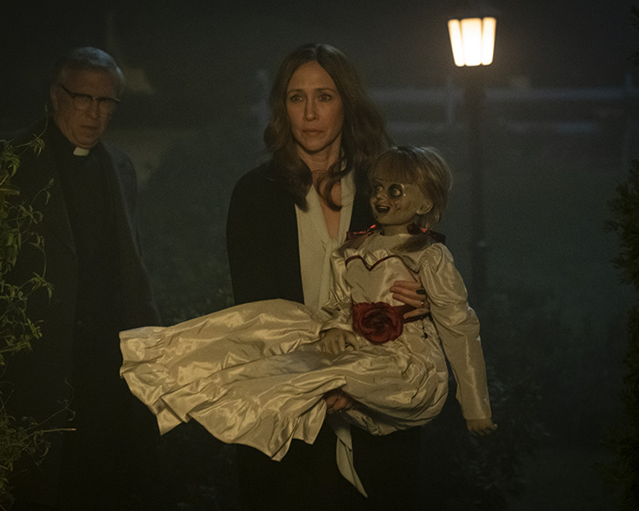 Woman holding haunted Annabelle doll at night with a priest in the background, highlighting the doll’s chilling history. Woman holding haunted Annabelle doll at night with a priest in the background, highlighting the doll’s chilling history.