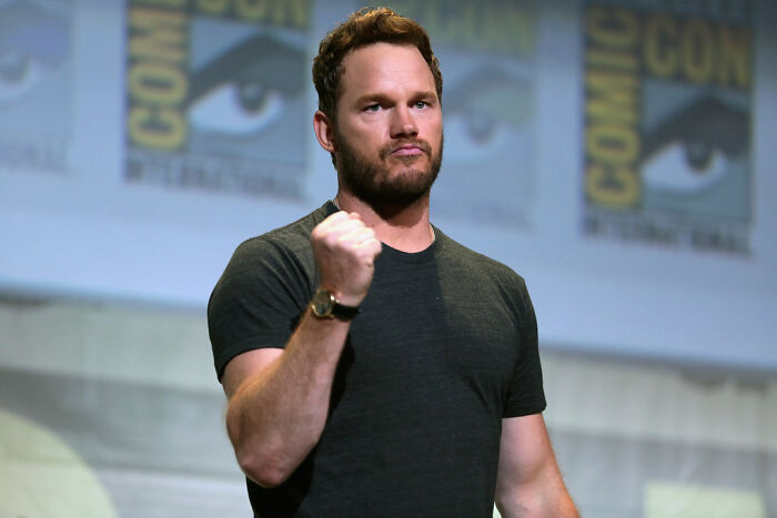 Man in a black t-shirt making a fist gesture on stage, representing celebrity gossip and juicy tea discussions.
