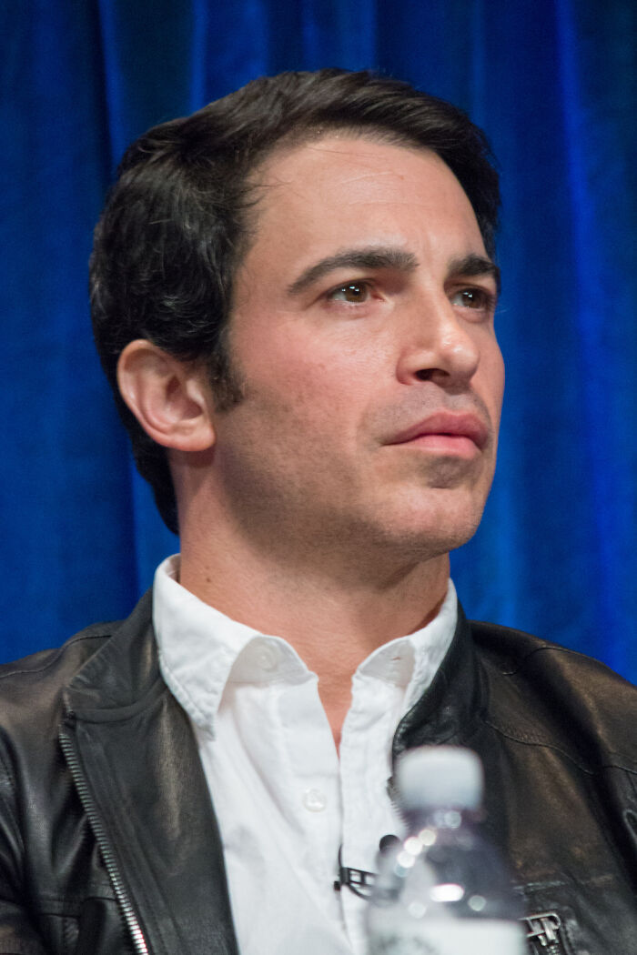 Man in a white shirt and leather jacket sitting in front of a blue curtain, themed around celebrity gossip and tea.