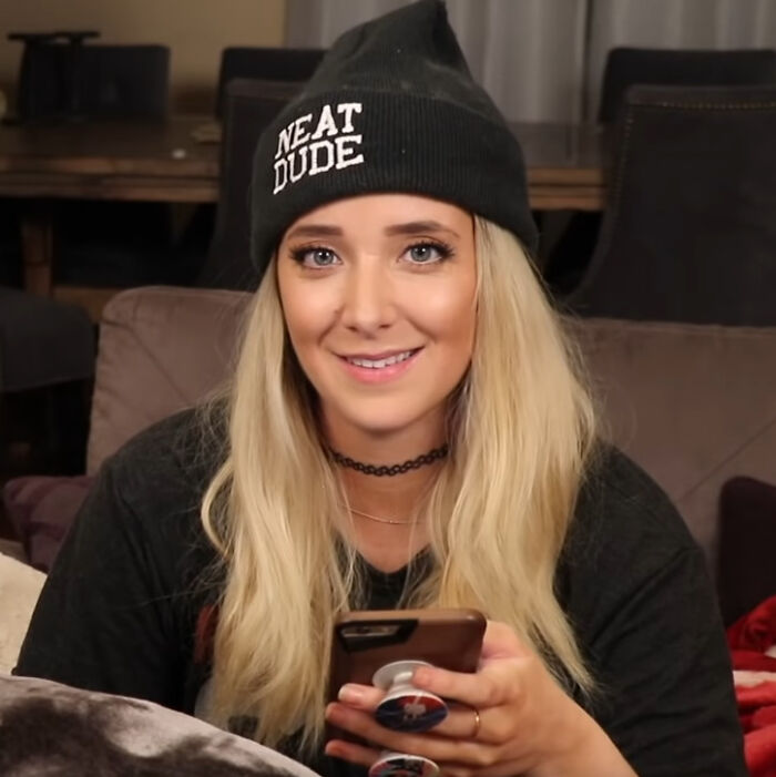 Young woman wearing a knit hat and holding a phone, sharing juicy celebrity gossip and tea in a casual setting.