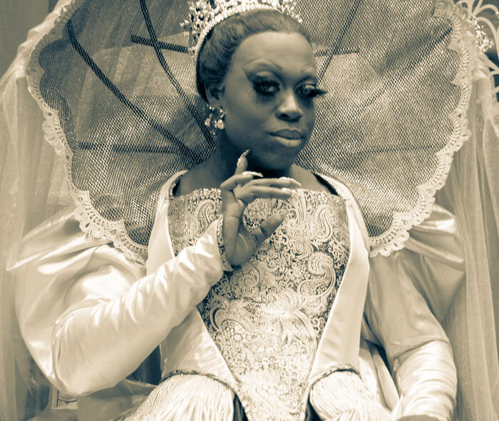 Person in ornate costume and crown, posing with hand gesture, representing celebrity gossip and the idea of spilling tea.