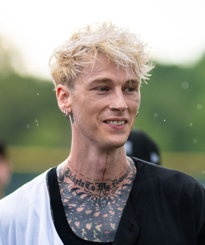 Young man with multiple earrings and neck tattoos, smiling outdoors, representing celebrity gossip and juicy tea topics.