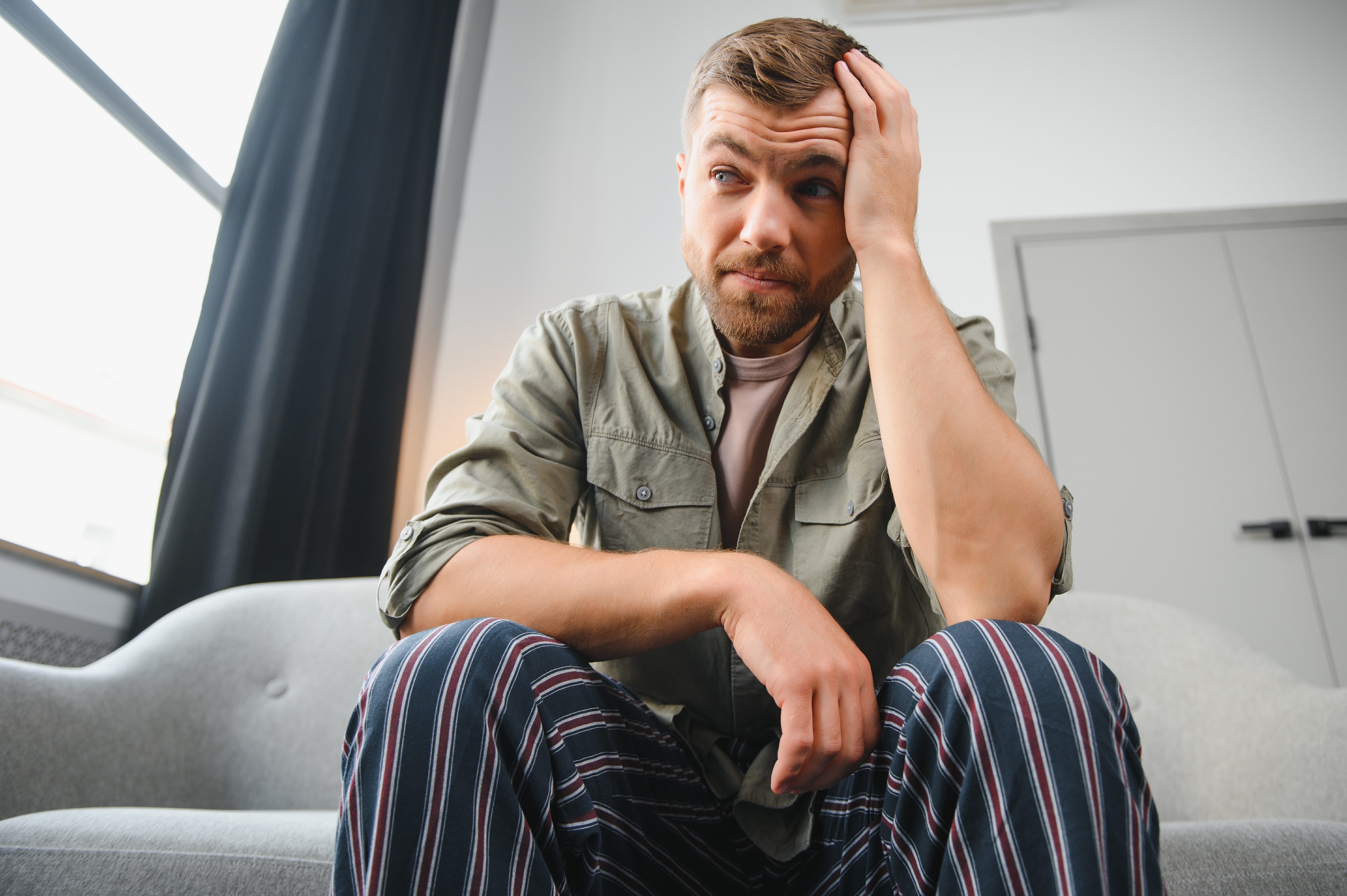A worried man sitting on a couch, wearing striped pants and an olive shirt, feeling embarrassed and stressed. A worried man sitting on a couch, wearing striped pants and an olive shirt, feeling embarrassed and stressed.