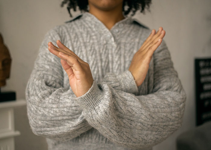 Person in gray sweater crossing arms in an X gesture, symbolizing rejection or denial of national stereotypes.