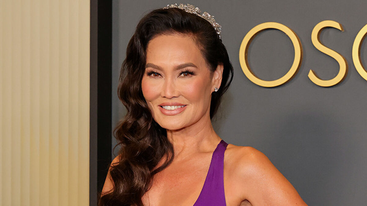 Tia Carrere, u201990s icon, wearing a plunging purple gown and tiara, smiling at an event with styled hair and elegant makeup.