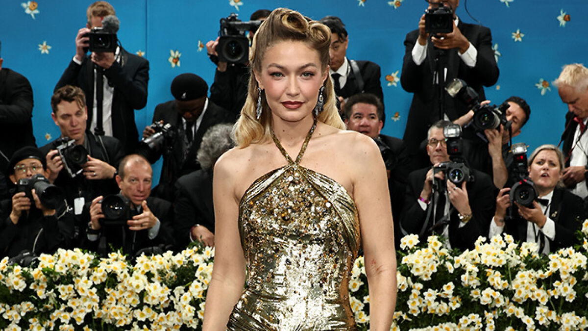 Gigi Hadid posing in a gold metallic dress at an event with photographers and floral decorations in the background.