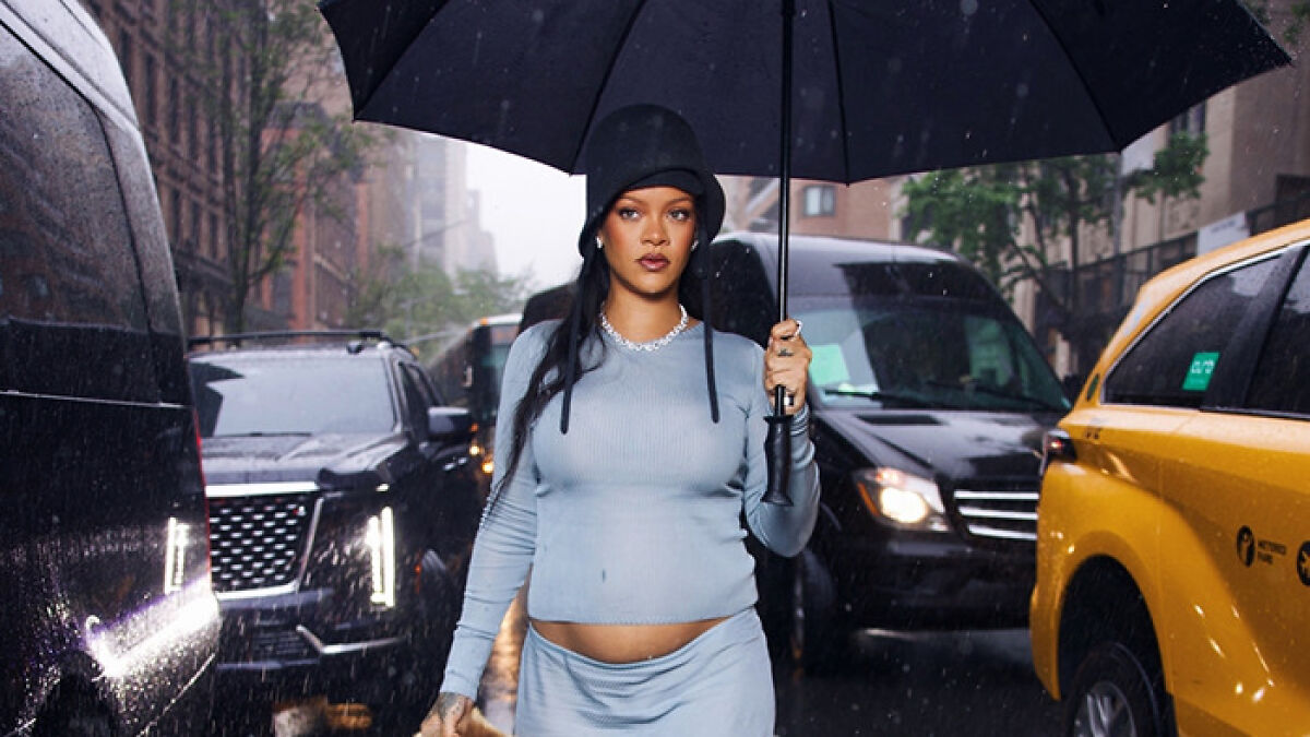 Rihanna showing her baby bump while holding an umbrella walking in the rain on a busy city street.