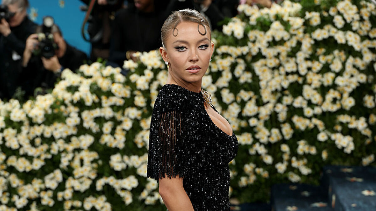 Sydney Sweeney in a black beaded outfit at the Met Gala, posing with a floral backdrop and photographers in the background