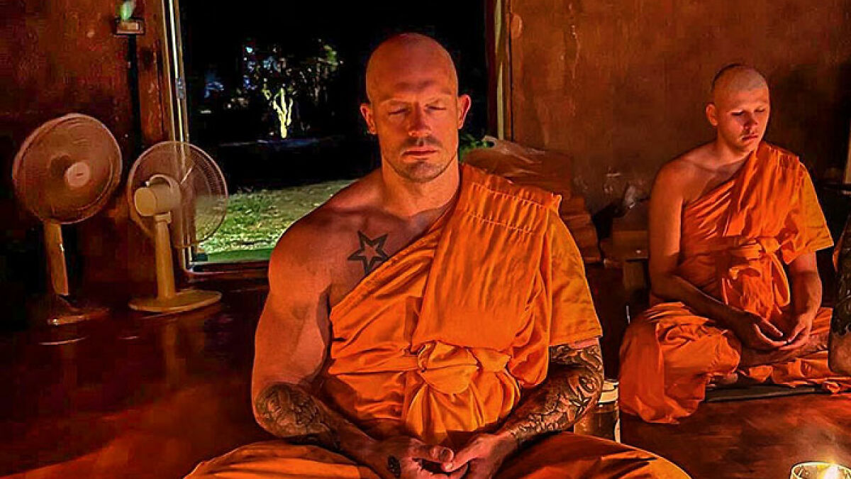 Former pole dancer in orange robes meditating calmly inside a dimly lit monk's room with another monk in the background.