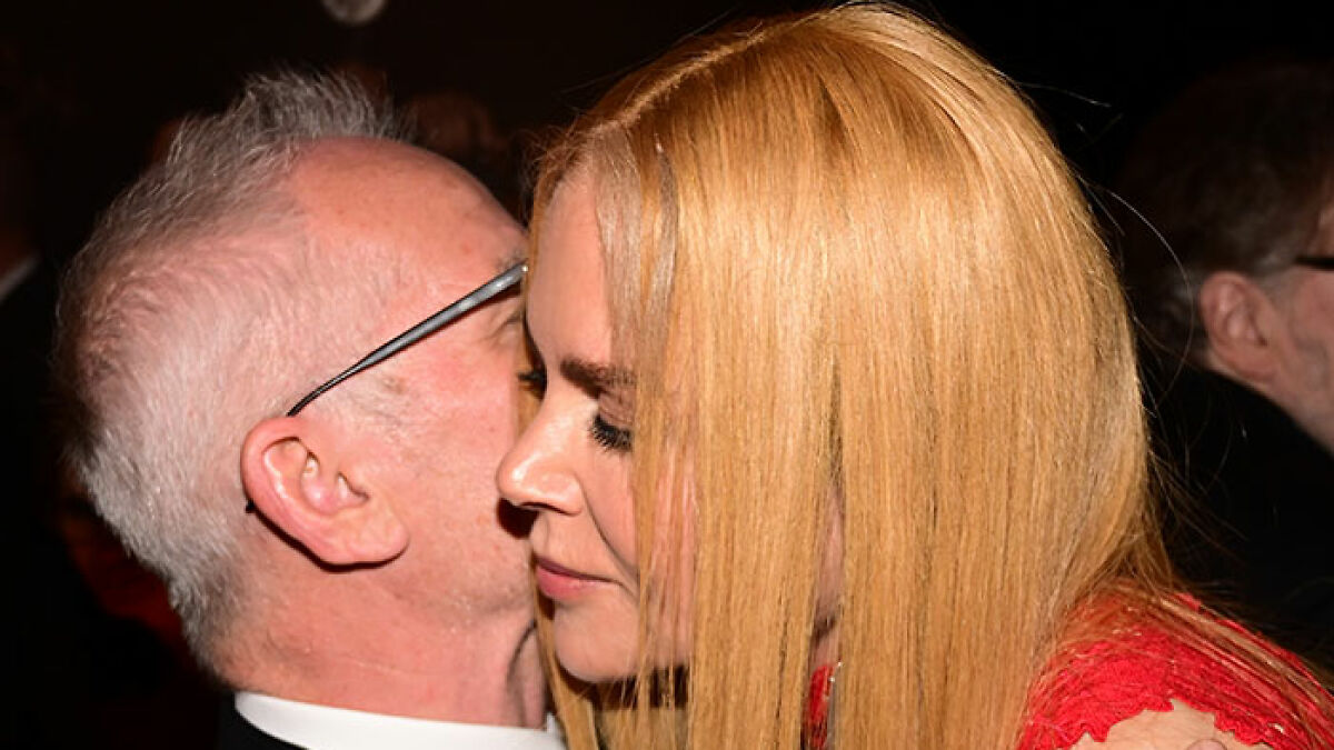 Nicole Kidman with blonde hair in a red dress sharing a close moment with a man wearing glasses at an event.