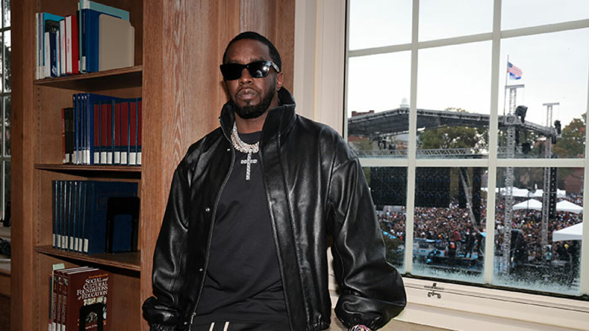 Diddy wearing sunglasses and a leather jacket indoors with a crowd and stage visible outside the window preparing for Freak Off.