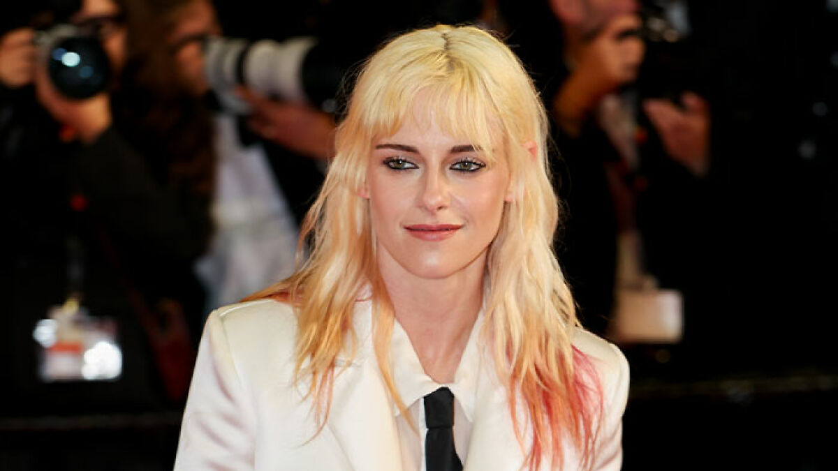 Kristen Stewart at a public event, smiling and wearing a white suit, with photographers in the background.