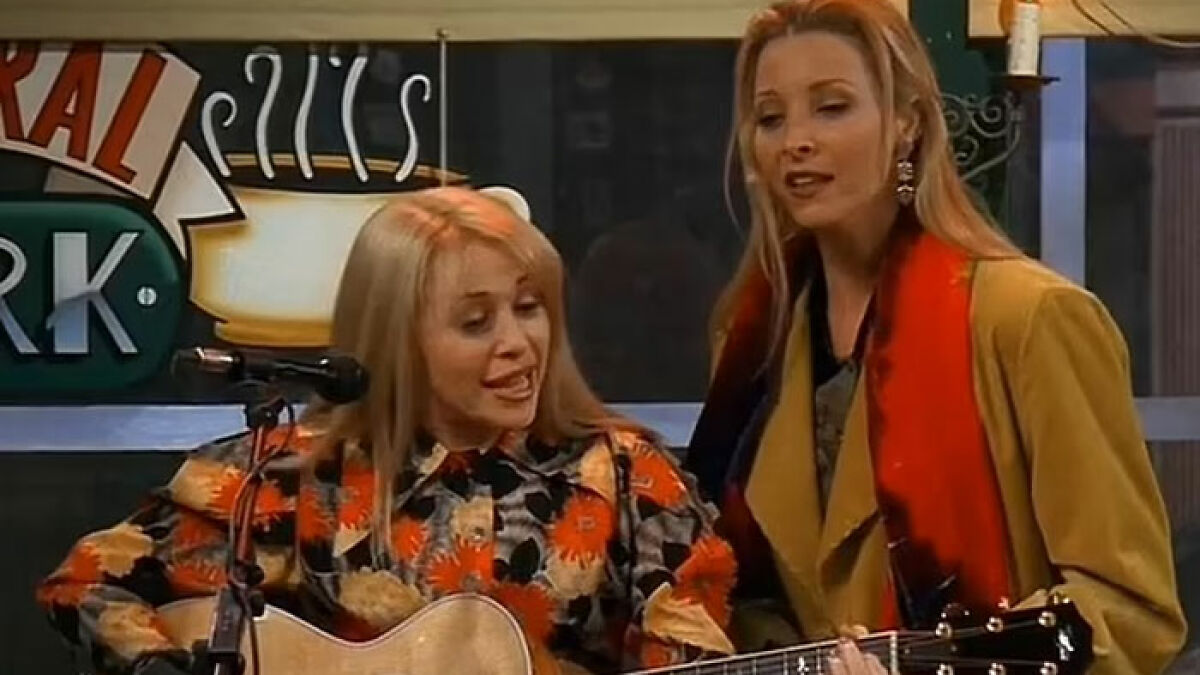 Friends star singing Smelly Cat with guitar, wearing floral and yellow outfits in a coffeehouse setting.