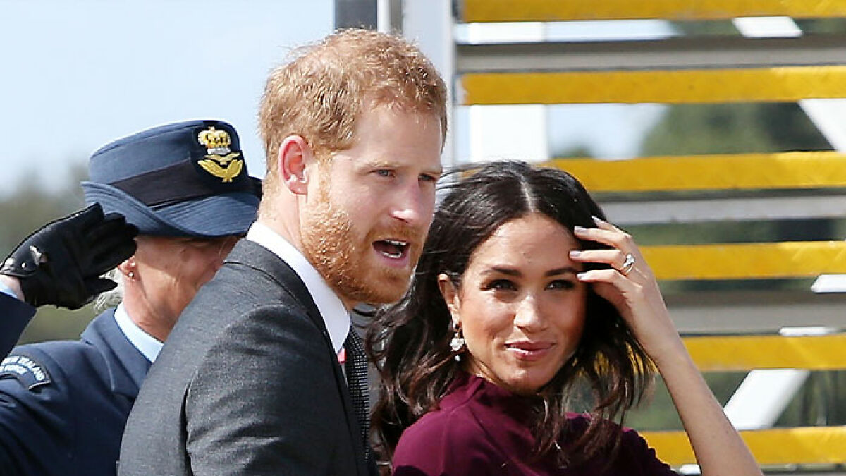 Meghan Markle and Prince Harry at an event outdoors, with Meghan touching her hair and Harry speaking urgently.