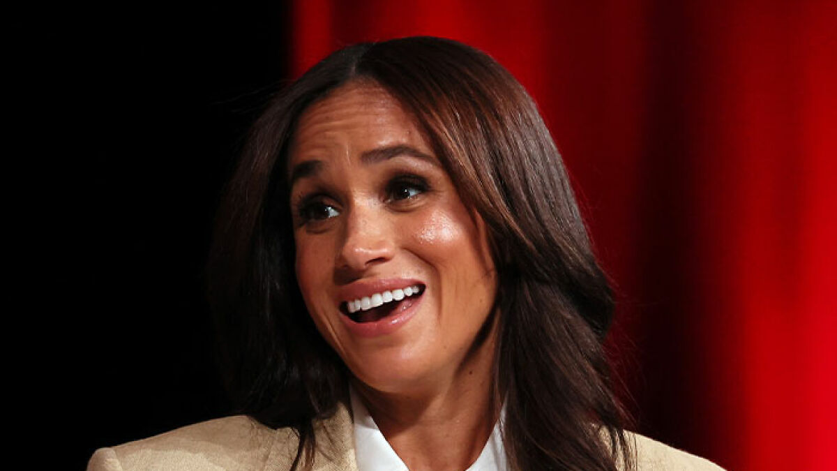 Meghan Markle smiling and talking during an event with a red curtain background, Vogue cover diva discussion.