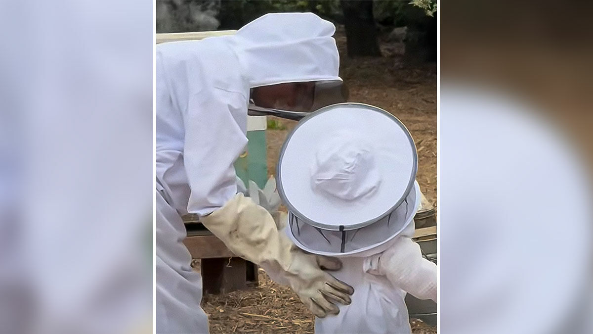 Meghan Markle and 3-year-old Lilibet in beekeeping suits outdoors, engaging with beekeeping equipment in natural setting
