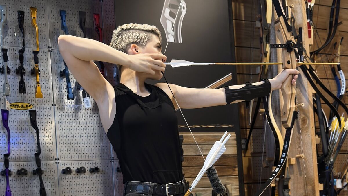 Trans archer aiming with bow in archery shop, surrounded by various bows and arrows on display.
