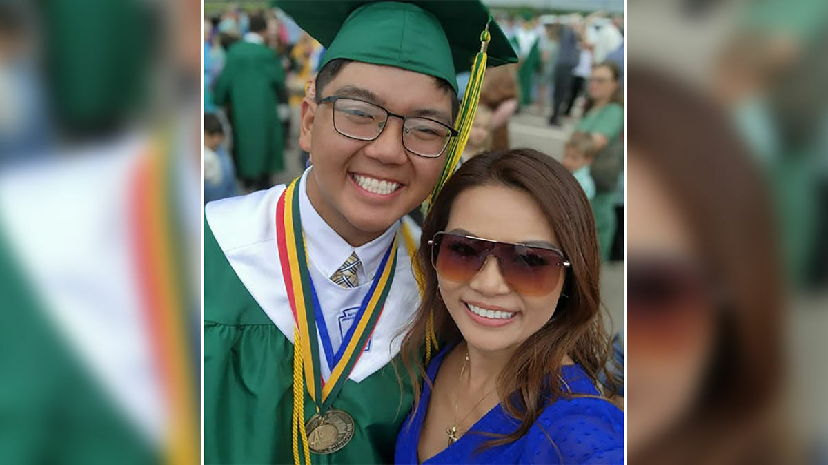 High school valedictorian in green cap and gown smiling with woman in sunglasses at graduation event.