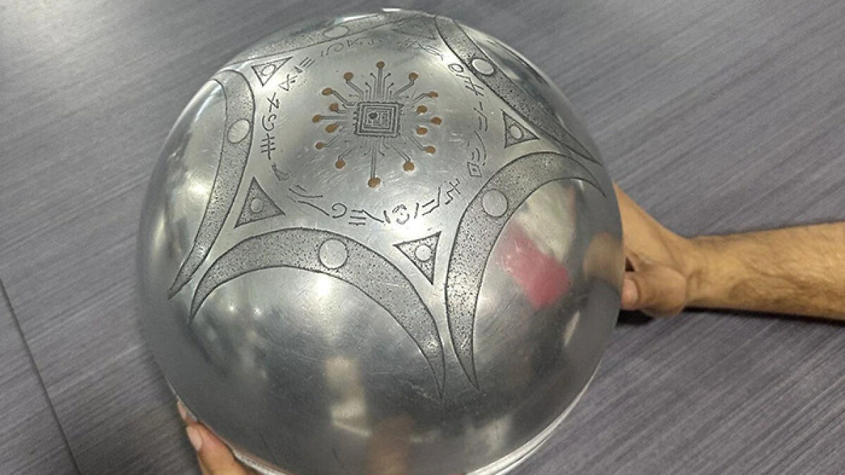 Metallic UFO with mysterious symbols and writing about origin of birth held by two hands on a gray surface.