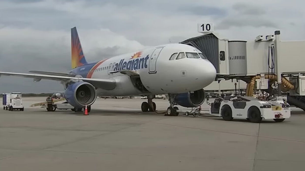 Allegiant airplane parked at gate with tow vehicle on airport tarmac under cloudy sky