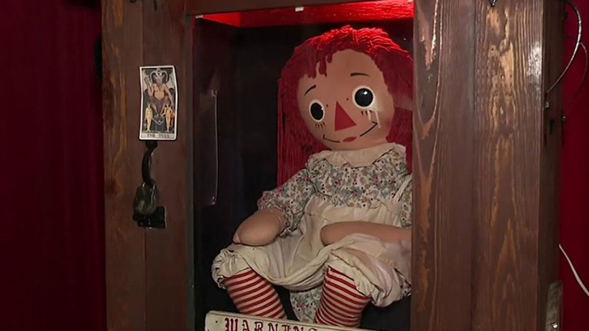Haunted Annabelle doll with red hair and striped stockings inside a wooden display case with warning sign and tarot card nearby