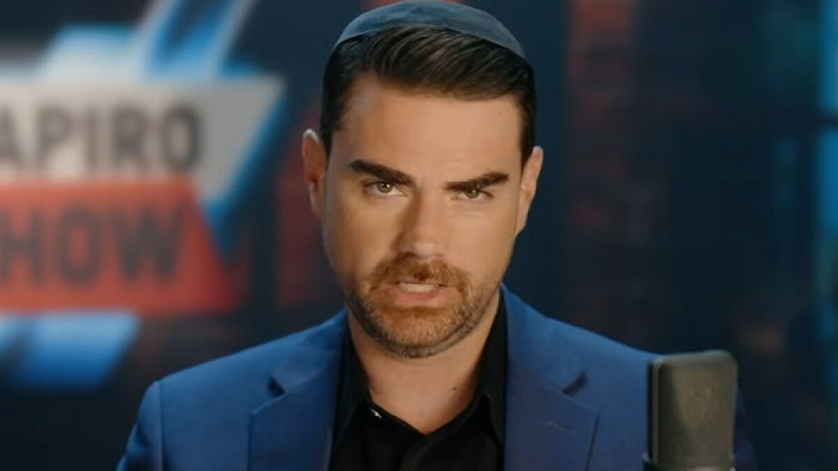 Ben Shapiro in a dark blue suit speaking into a microphone during a show with a blurred background.