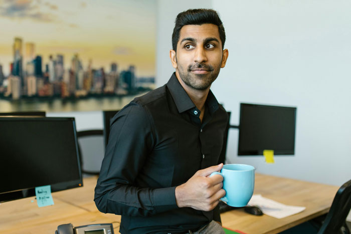 Man in office holding a blue mug, looking away thoughtfully, with computer monitors in the background.