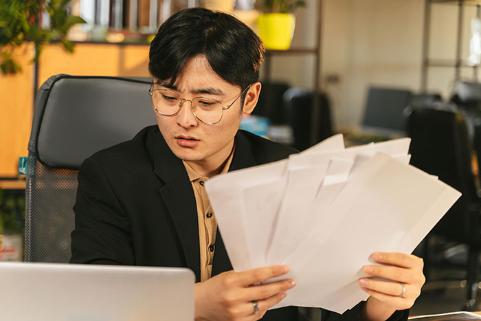 Man in glasses looking stressed while holding papers at office desk, depicting helicopter mom calls to son's boss scenario. Man in glasses looking stressed while holding papers at office desk, depicting helicopter mom calls to son's boss scenario.