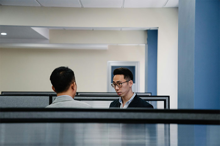 Two men in an office cubicle having a serious conversation, illustrating a helicopter mom calling 30YO son’s boss. Two men in an office cubicle having a serious conversation, illustrating a helicopter mom calling 30YO son’s boss.