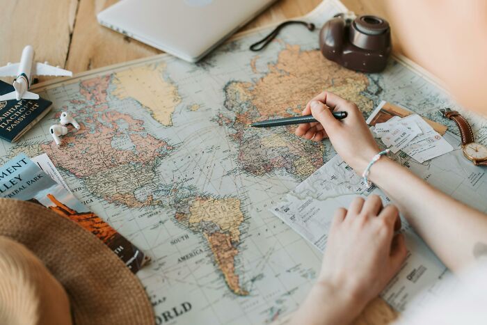 Hands marking a world map during family trip planning, with travel tickets, camera, and passport nearby. Hands marking a world map during family trip planning, with travel tickets, camera, and passport nearby.