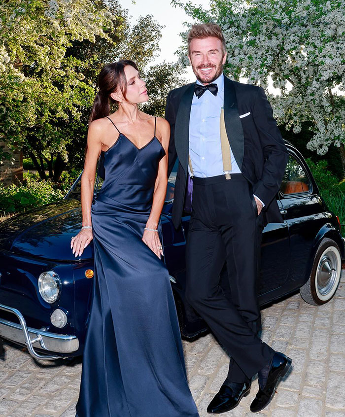David Beckham and Victoria Beckham dressed formally posing by a vintage car at a 50th birthday party event. David Beckham and Victoria Beckham dressed formally posing by a vintage car at a 50th birthday party event.