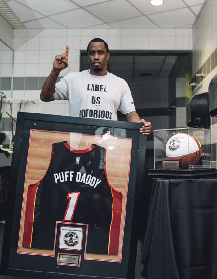 Man holding framed Puff Daddy basketball jersey and basketball display case, related to Diddy trial Miami mansion raid items. Man holding framed Puff Daddy basketball jersey and basketball display case, related to Diddy trial Miami mansion raid items.