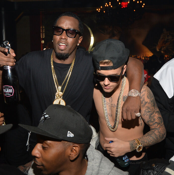 Diddy with gold chains and sunglasses holding a champagne bottle, rapper shirtless wearing chains and a cap in a club setting