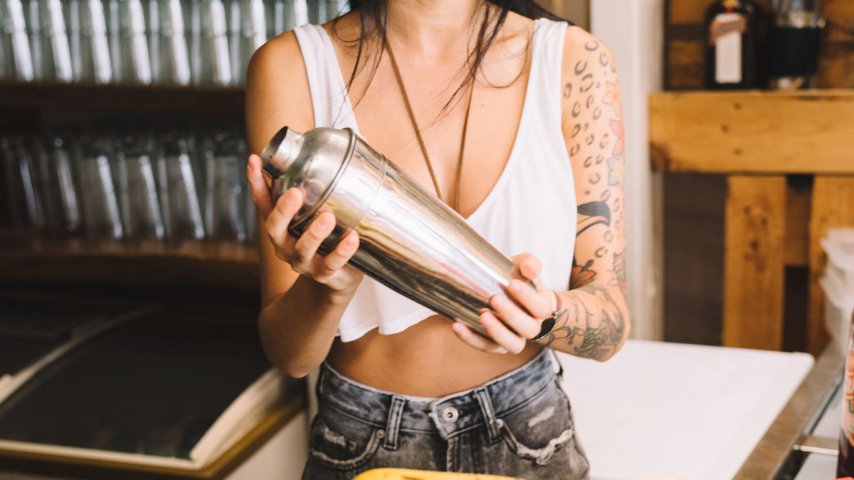 Student shaking a cocktail mixer at a bar, wearing a white crop top and showing tattooed arms in a casual setting