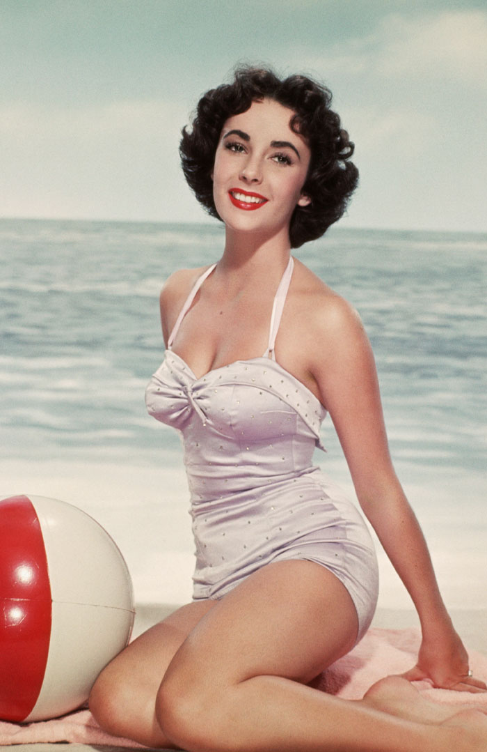 Elizabeth Taylor smiling by the ocean in a vintage swimsuit, her eyes capturing mystery in pop culture.