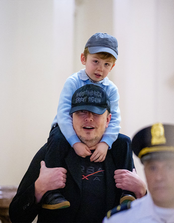 Elon Musk carrying a child on his shoulders, wearing a black cap, raising concerns after glitching and black eye appearance. Elon Musk carrying a child on his shoulders, wearing a black cap, raising concerns after glitching and black eye appearance.