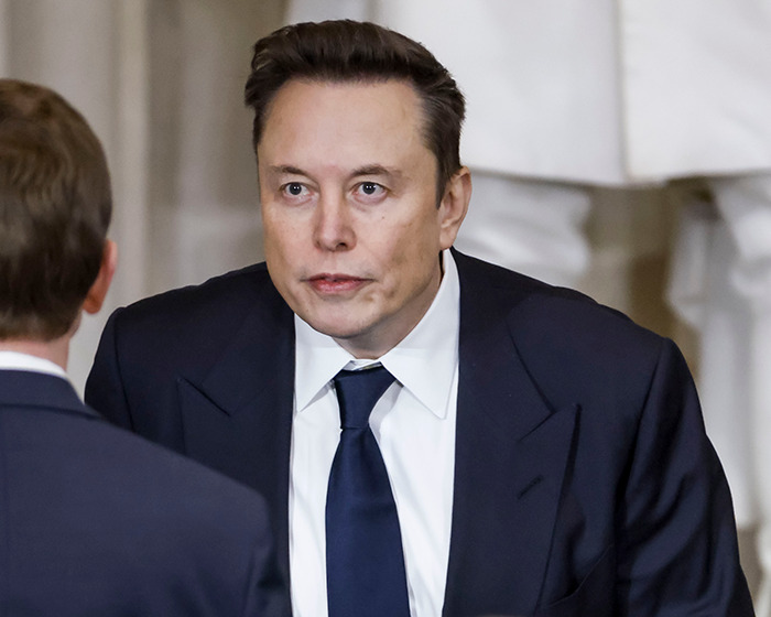 Elon Musk in a dark suit and tie engaged in conversation, facing another person in a formal indoor setting. Elon Musk in a dark suit and tie engaged in conversation, facing another person in a formal indoor setting.