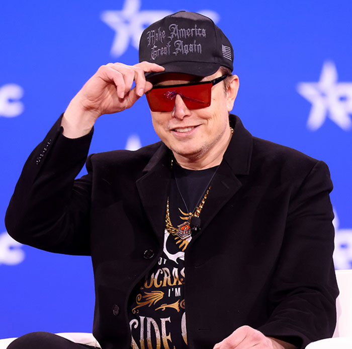 Elon Musk wearing sunglasses and a cap, smiling during a public speaking event with a blue background. Elon Musk wearing sunglasses and a cap, smiling during a public speaking event with a blue background.