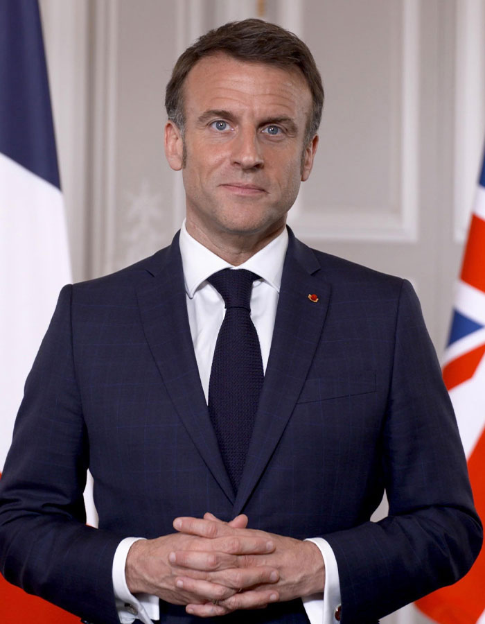 French President Emmanuel Macron in a formal suit standing between French and British flags during a speech.