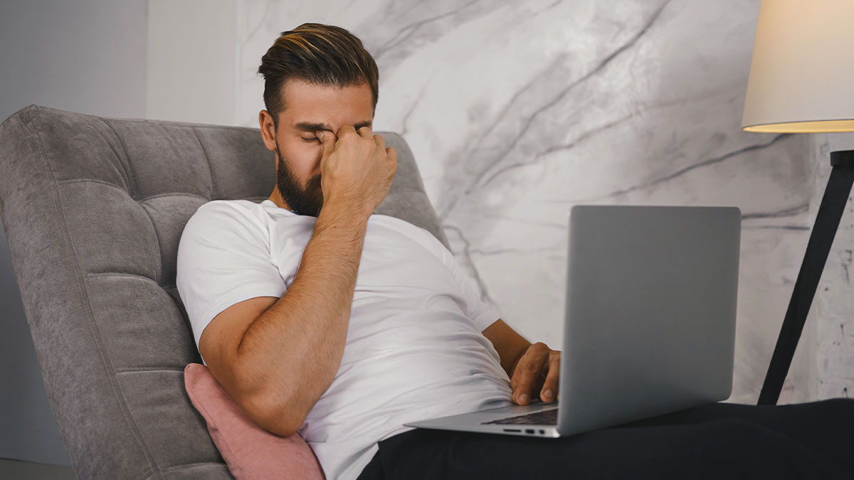 Man in a white shirt sitting on a gray chair, looking stressed while using a laptop at home during wellness checks.