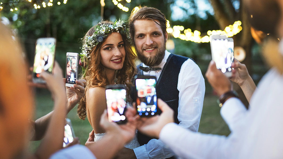 Bride and groom posing for photos at outdoor wedding surrounded by guests capturing moments on smartphones, wedding revenge controversy.