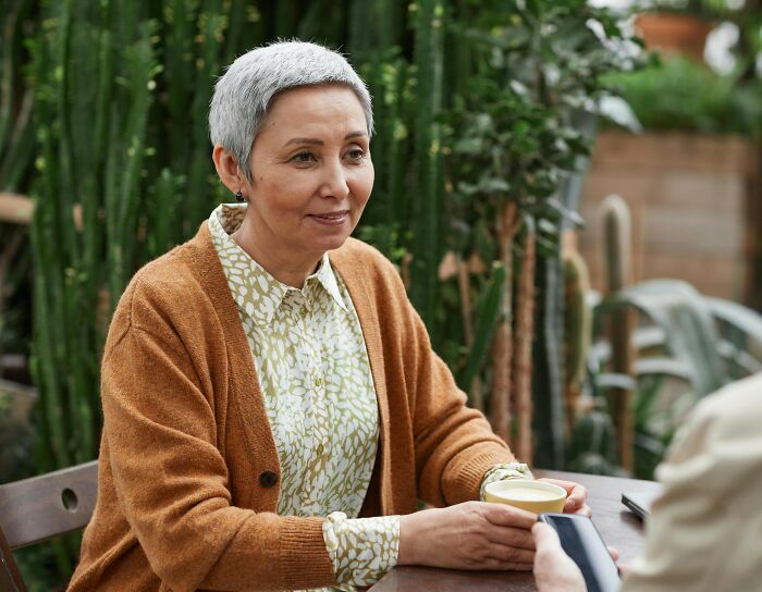 Older woman with short gray hair sitting at a table holding a cup, reflecting feelings of jealous grandparents and generosity conflict. Older woman with short gray hair sitting at a table holding a cup, reflecting feelings of jealous grandparents and generosity conflict.