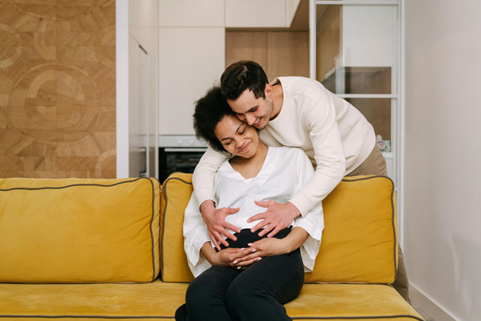 Couple embracing on yellow couch as pregnant woman sits smiling, highlighting wedding venue booking and family surprises. Couple embracing on yellow couch as pregnant woman sits smiling, highlighting wedding venue booking and family surprises.