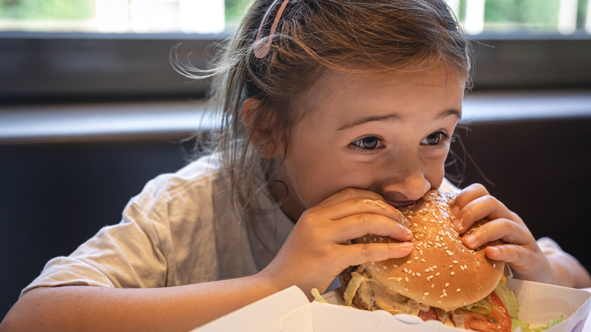 Young girl enjoying a large cheeseburger indoors, highlighting junk food feeding and indulgence with nieces context.