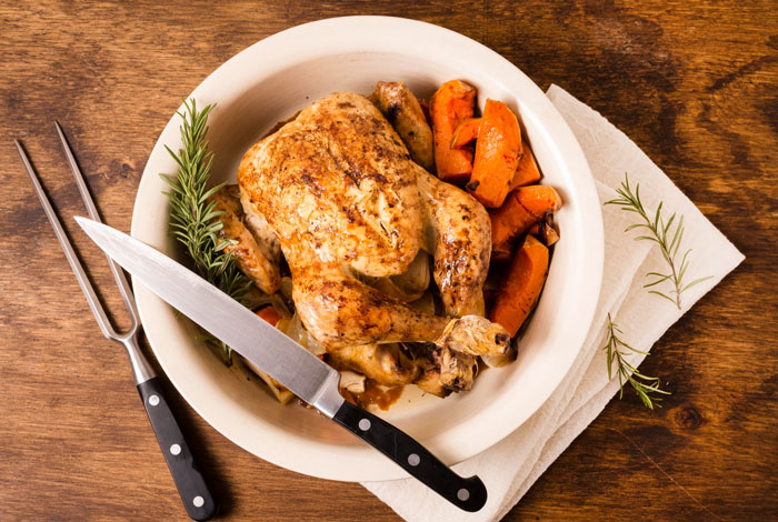 Roasted chicken with carrots on a white plate beside carving knife and fork on wooden table with herbs Roasted chicken with carrots on a white plate beside carving knife and fork on wooden table with herbs