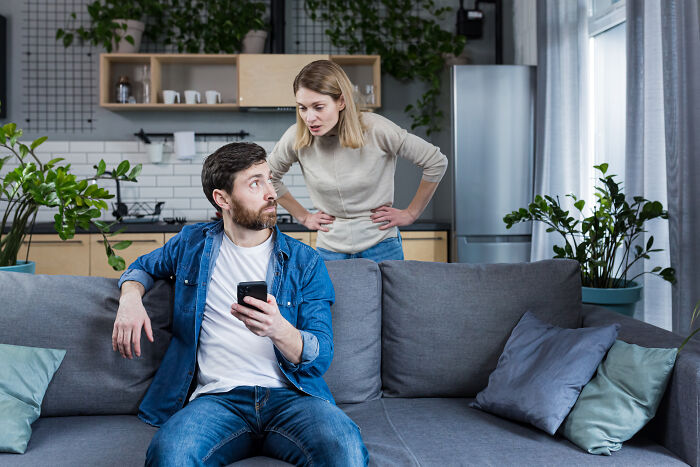Man sitting on couch holding phone while woman stands behind him angrily confronting him about turning his back on female friends.